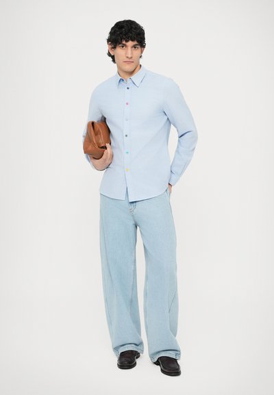 Cămașă albastru deschis cu nasturi colorați, blugi deschiși la culoare, croială lejeră, ținând o geantă de mână de piele maro, pantofi negri.