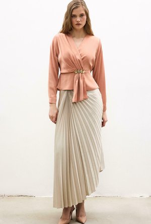 Kobieta w brzoskwiniowej bluzce zapinanej kopertowo z złotymi guzikami oraz w beżowej plisowanej spódnicy midi z wysokim stanem, stojąca na tle białej ściany.