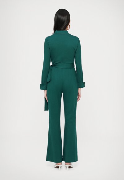 Zöld átugró ruházat hosszú ujjakkal, öves derékkal és bővülő szárakkal. Simább anyag és gombos mandzsetták; fehér magassarkúval párosítva.