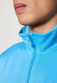 Ljusblå zip-jacka med hög krage, mjuk textur och smidig dragkedja. Har minimalt med sömmar och en strömlinjeformad design.