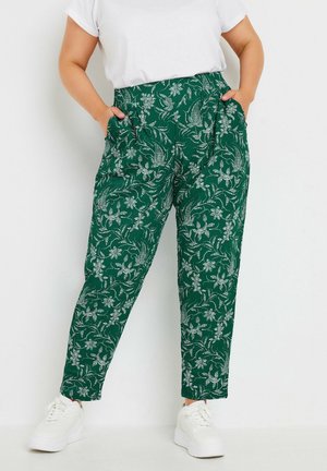 Personne portant un pantalon vert à motifs floraux avec des poches et des baskets blanches, debout les mains dans les poches sur un fond blanc.