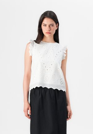 Model poses in a white embroidered top with ruffled sleeves, paired with a black skirt against a plain white background.