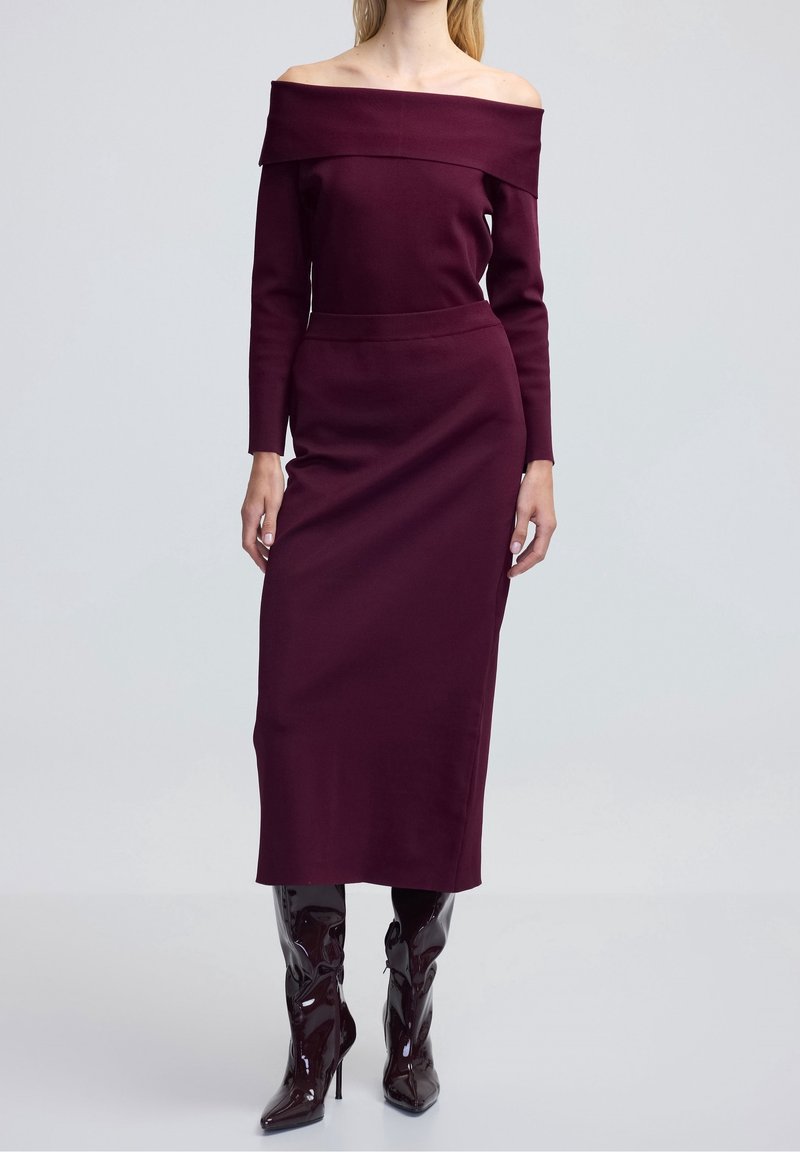 Robe bordeaux à épaules dénudées avec des manches longues, silhouette ajustée et longueur mi-mollet, associée à des bottes noires laquées hautes.