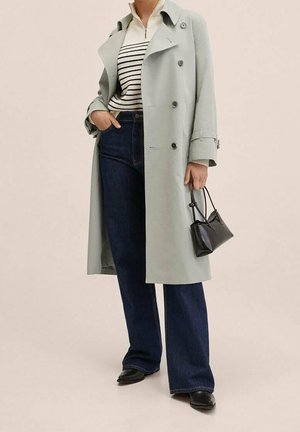 Femme portant un trench-coat gris clair, un pull zippé rayé, un jean bleu foncé, des chaussures noires, tenant un petit sac à main noir.