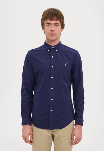 Camisa azul-marinho de botão em algodão com colarinho pontudo, apresentando pequenos botões brancos e um detalhe de logótipo dourado no lado esquerdo do peito.