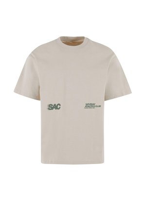 T-shirt a maniche corte di colore beige chiaro realizzato in cotone. Presenta il logo "SAC" e il testo "SATURDAY ATHLETICS CLUB" in verde sulla parte anteriore.