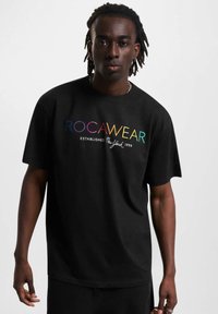 Juodas trumpas rankovės marškinėlis su daugiaspalviu "ROCAWEAR" logotipu ir užrašu "Įkurta Niujorke 1999". Lygus medvilnės audinys, laisvas siluetas.