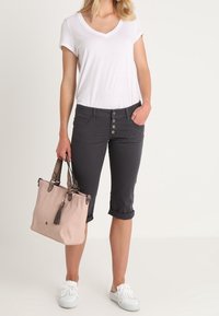 Pantalons gris courts avec une braguette à boutons, associés à un t-shirt blanc en v et des mocassins blancs. Un sac à main rose avec des accents marron est tenu dans une main.