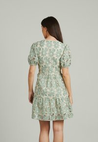 Mujer con cabello largo y oscuro que lleva un vestido corto de mangas abullonadas con una superposición de encaje floral verde, de espaldas contra un fondo liso.