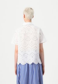White embroidered blouse with short sleeves, featuring cut-out patterns and a scalloped hem, paired with a blue and white striped skirt.