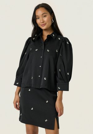 Eine Frau trägt eine schwarze Bluse mit Knopfleiste und einen passenden Rock mit silbernen Paillettenverzierungen. Sie steht vor einem einfarbigen hellen Hintergrund.