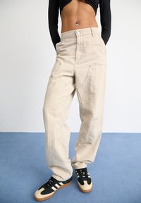 Beige oversized broek van canvas met grote zakken, knoopsluiting en subtiele stikdetails, gecombineerd met zwart en crèmekleurige sneakers.