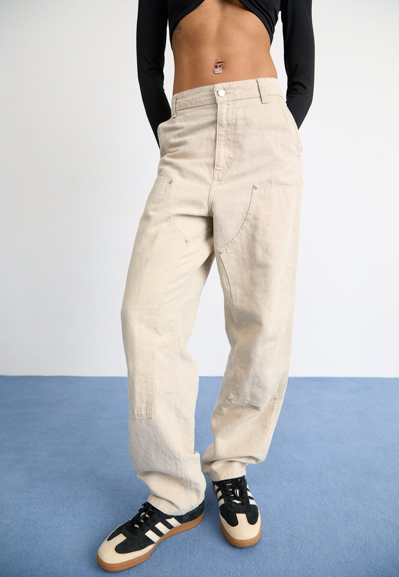 Beige oversized broek van canvas met grote zakken, knoopsluiting en subtiele stikdetails, gecombineerd met zwart en crèmekleurige sneakers.