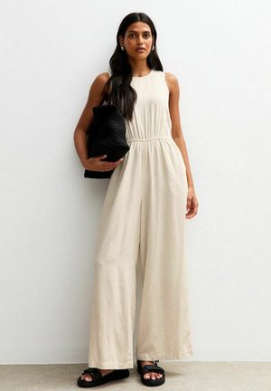 Beige mouwloze jumpsuit met een aansluitende taille, vloeiende wijde pijpen, gecombineerd met een zwarte textuur tas en zwarte sandalen. Vloeiende stoffen textuur.