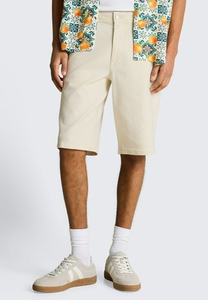 Mann trägt beige knielange Shorts, weiße Socken, beige Sneakers und ein gemustertes Hemd mit Orangen- und grünen Designs, steht vor einem einfarbigen Hintergrund.