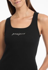 Black ribbed tank top featuring a scoop neckline and embroidered "guessjeans" logo in white, highlighting a slim fit design.