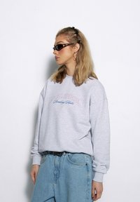 Szary, oversize'owy sweatshirt z haftowanym napisem "Lokalni Bohaterowie Łamią Zasady". Noszony z jasnoniebieskimi, luźnymi dżinsami.