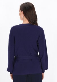 Pull en maille côtelée marine avec une taille ceinturée, des manches larges et un col rond, présentant un tissu texturé et sans motifs visibles.