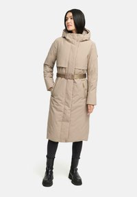Beige polstret jakke med lynlås, hætte og bælte. Har en mellemlang design og sidelommer. Båret over mørke tætsiddende bukser og støvler.