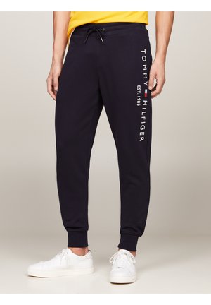 Man in marineblauwe Tommy Hilfiger trainingsbroek met logo-tekst en witte sneakers, staand tegen een neutrale achtergrond.