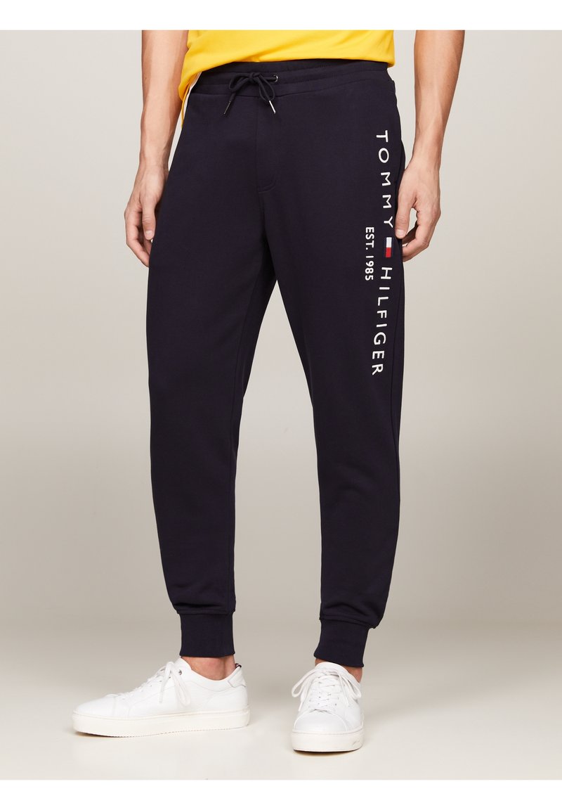 Man som bär marinblå Tommy Hilfiger joggingsbyxor med logotext och vita sneakers, står mot en neutral bakgrund.