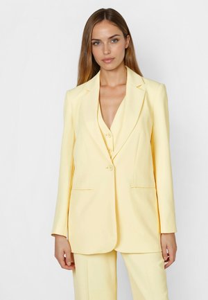 Frau mit langen, glatten Haaren, die einen hellgelben, taillierten Blazer und passende Hose trägt, vor einem weißen, schlichten Hintergrund stehend.