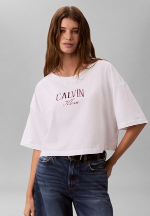 Jeune femme aux longs cheveux bruns porte un t-shirt blanc court Calvin Klein et un jean bleu sur fond gris.