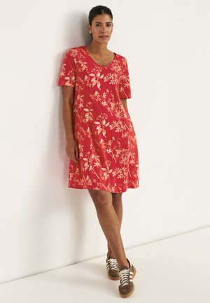 Kvinde iført en rød blomsterprintet kjole til knæene med korte ærmer, stående op ad en hvid væg, brune sneakers med hvide snørebånd.