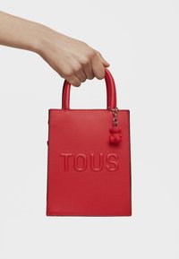 Bolso de mano de piel roja con texto en relieve "TOUS", asas cortas y un pequeño charm de oso rojo colgando de una cadena de tono plateado.