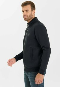 Uomo con i capelli corti e la barba, che indossa una giacca nera con zip e jeans blu, in piedi contro uno sfondo bianco semplice.