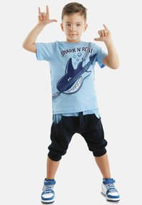 Ljusblå t-shirt med en hajgrafik och texten "SHARK 'N' ROLL", i kombination med svarta shorts och blå sportskor.