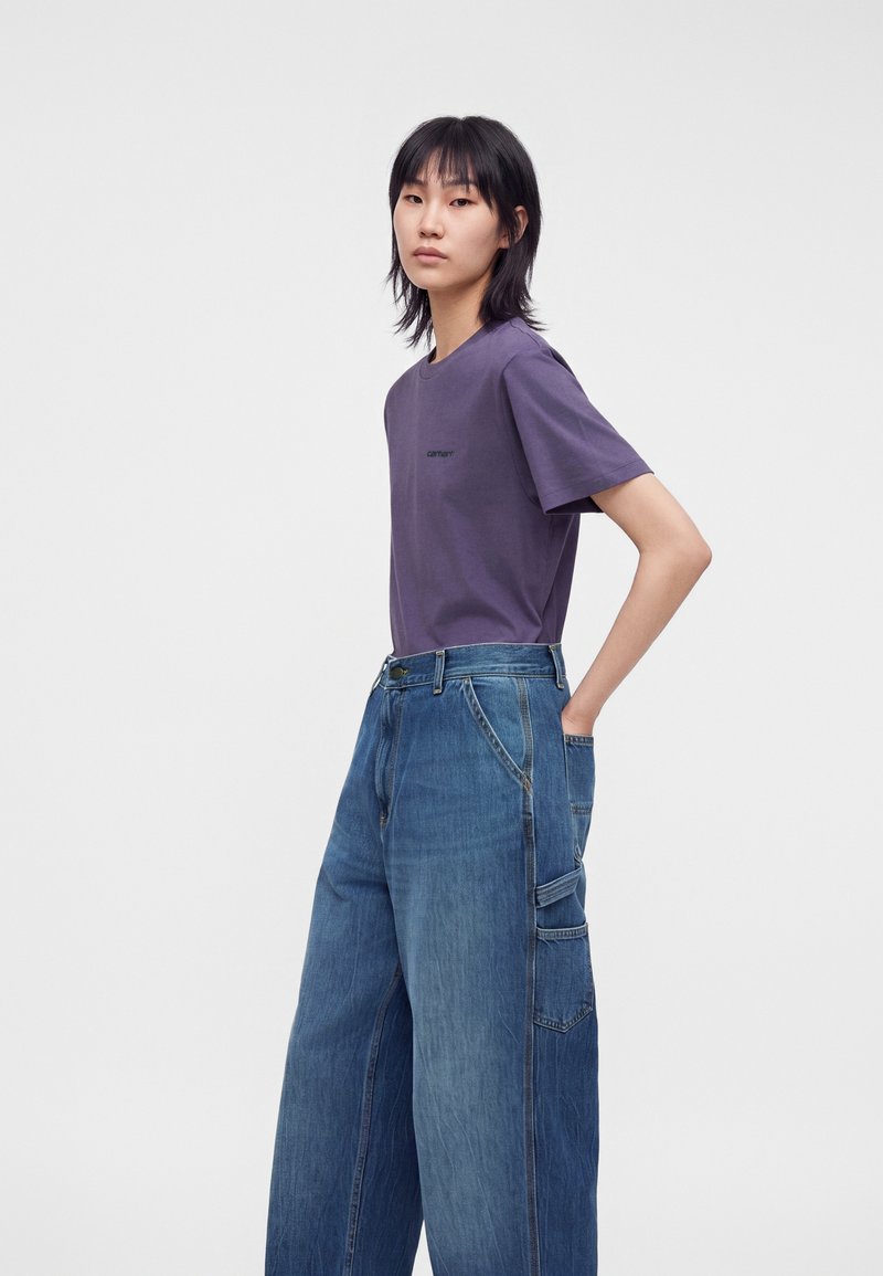 Young person with black hair wears a purple t-shirt and wide-leg blue jeans with cargo pockets against a plain light gray background.