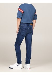 Ciemnoniebieskie dżinsy o wąskim kroju, z tylnymi kieszeniami i brązową skórzaną metką. Zestawione z niebieską koszulą i białymi trampkami.