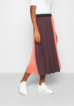 Femme portant une jupe midi plissée avec des panneaux violet foncé et pêche, des baskets blanches et un haut blanc à manches courtes sur un fond uni.