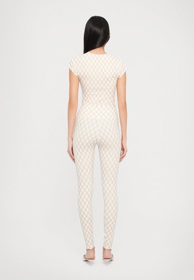 Strak zittende jumpsuit in beige met een geometrisch patroon. Heeft kapmouwen en een gladde textuur, gecombineerd met hoge muiltjes.