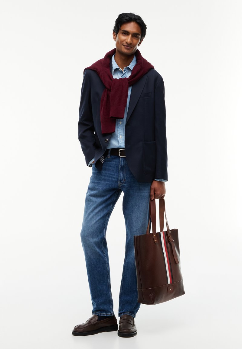 Jeune homme portant un blazer bleu marine, une chemise bleu clair, un jean bleu, avec un pull bordeaux sur les épaules, tenant un sac cabas marron avec un détail rayé.