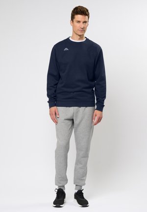 Felpa blu navy con collo a giro, polsini e orlo a coste; abbinata a pantaloni jogger grigi chiari e scarpe da ginnastica nere.