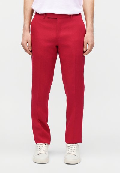 Άτομο που φορά φωτεινά κόκκινα στενά παντελόνια και λευκά αθλητικά παπούτσια στέκεται μπροστά σε ένα απλό φωτεινό φόντο.