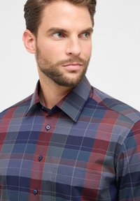 Homme barbu regardant vers la droite, portant une chemise à carreaux boutonnée dans des tons de bleu, rouge et gris, sur un fond clair uni.