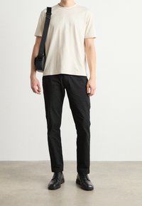 T-shirt di cotone beige con logo in rilievo, abbinata a pantaloni neri aderenti e scarpe in pelle nere. Una borsa a tracolla pende dalla spalla sinistra.