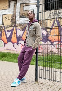 Giacca di jeans grigia chiara, felpa viola e pantaloni della tuta viola. Le scarpe hanno dettagli color turchese e rosa. In piedi vicino a un muro di graffiti.