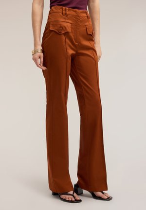 Femme portant un pantalon taille haute marron rouille avec poches à rabat devant et des sandales noires à talons blocs, debout devant un fond uni.