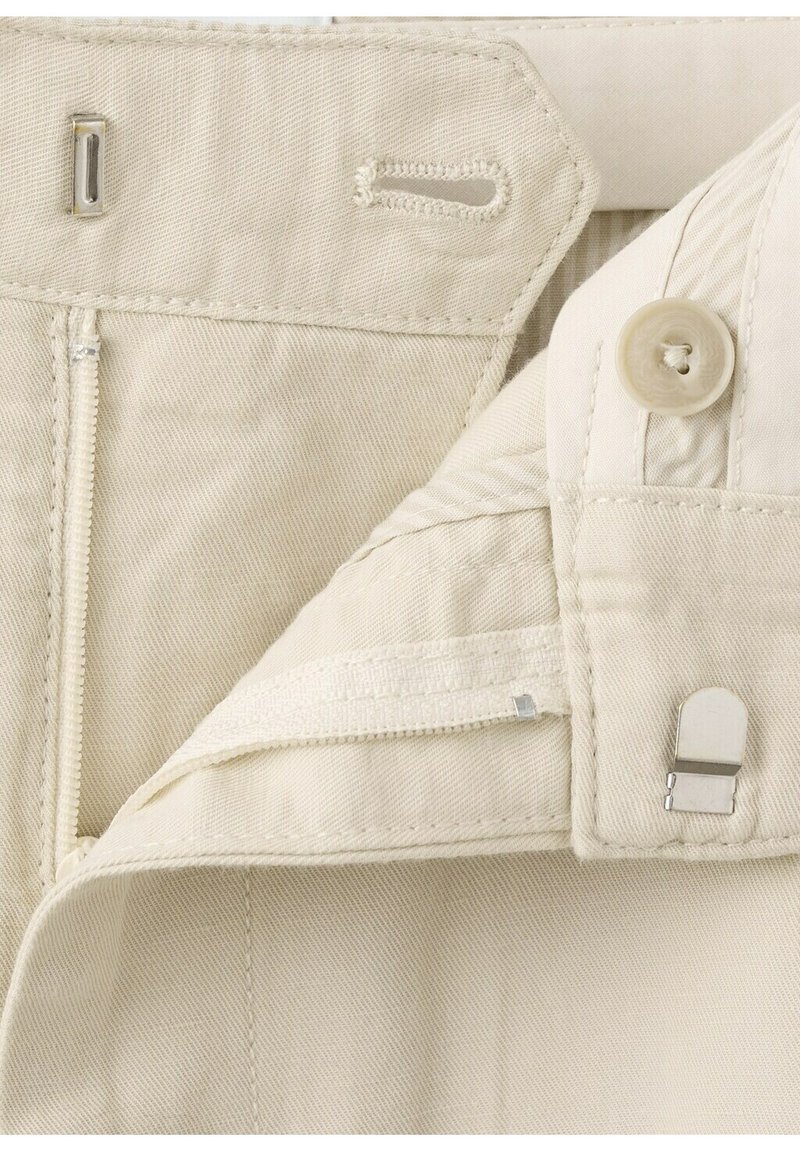 Tissu beige avec une fermeture éclair métallique, un bouton, une fermeture à agrafe avec œillet, et une boutonnière cousue, montrant les composants de fixation en détail.