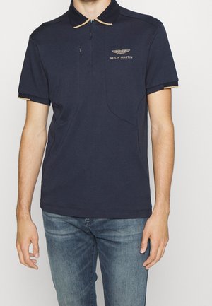 Homme portant un polo Aston Martin bleu marine avec un liseré beige au col et aux manches, associé à un jean bleu, debout devant un fond uni.