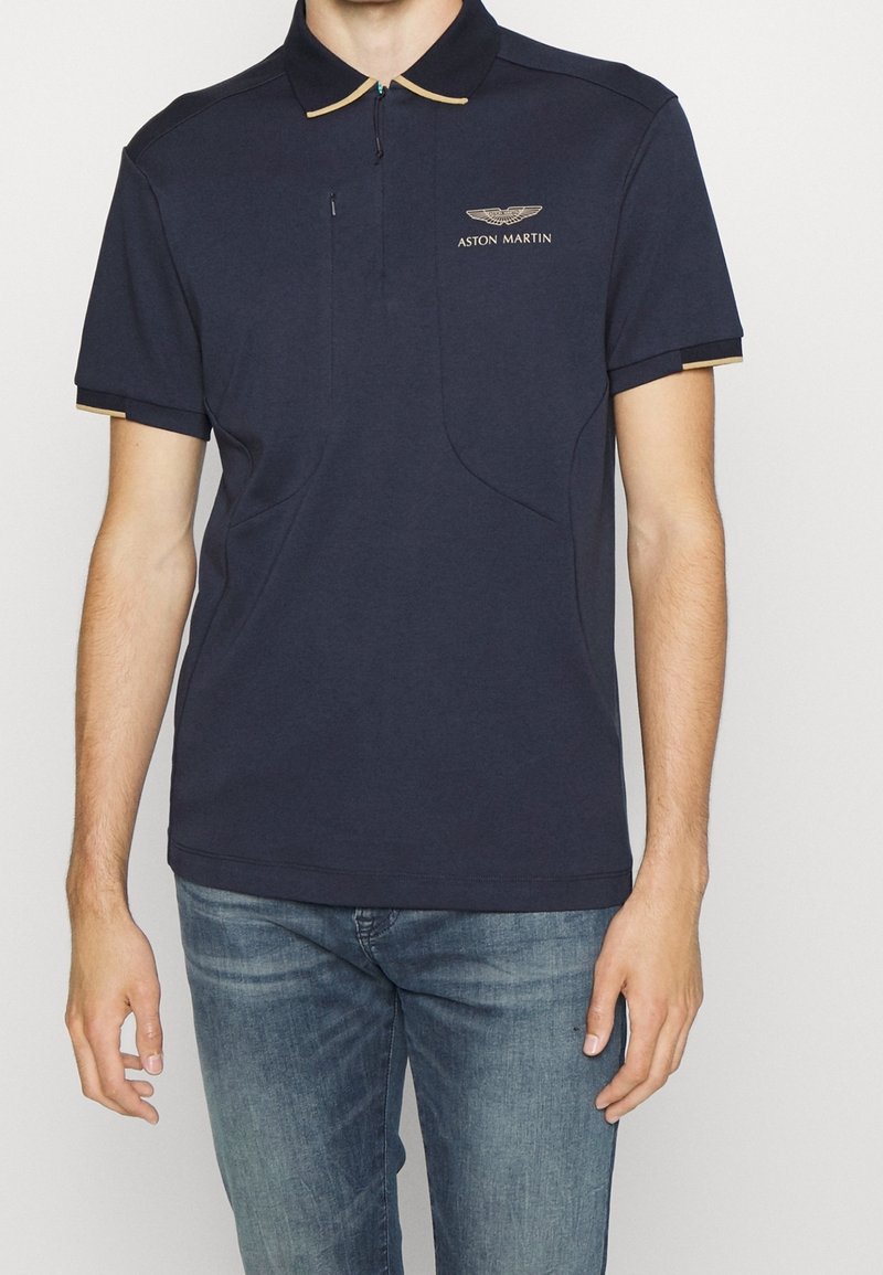 Homme portant un polo Aston Martin bleu marine avec un liseré beige au col et aux manches, associé à un jean bleu, debout devant un fond uni.