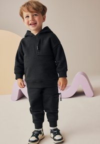 Sweat-shirt à capuche noire avec une poche avant, associé à un pantalon de jogging fuselé assorti avec une poche zippée sur le côté, porté avec des baskets noir et blanc.
