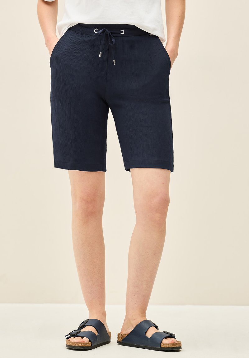 Shorts bleu marine arrivant aux genoux en tissu texturé avec une taille élastique et un cordon de serrage. Associés à des sandales bleu marine à enfiler avec des semelles en liège.