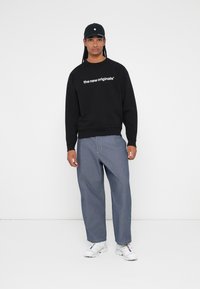 Sweatshirt preto com texto branco, calças de ganga azul de corte largo e sapatilhas brancas desportivas. O modelo usa um boné preto e está posicionado contra um fundo simples.