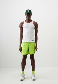 Tank top in white with orange trim, paired with bright green shorts, green socks, and white sneakers. Model wearing a green cap with a number 9.
