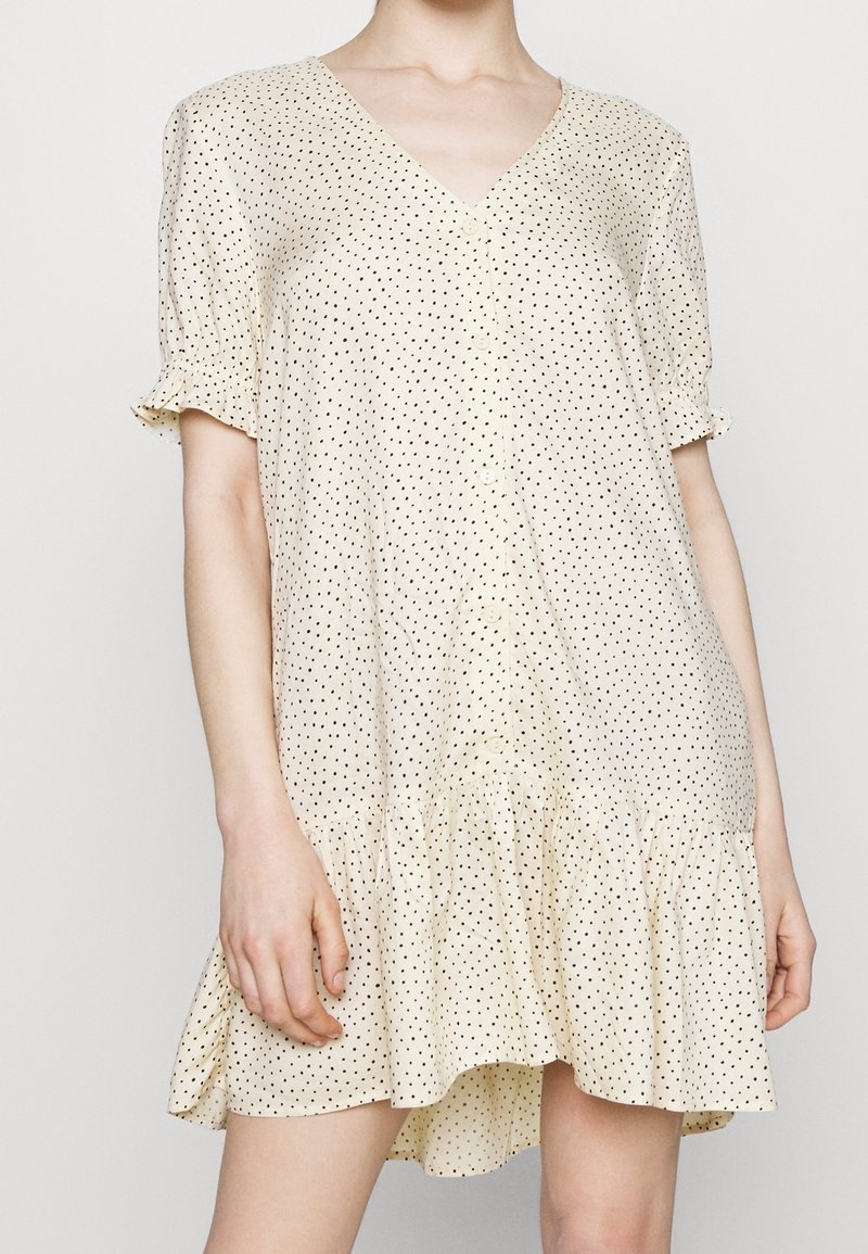Femme portant une robe ample crème à manches courtes avec des pois noirs, boutonnée devant et ourlet volanté, debout devant un fond uni.
